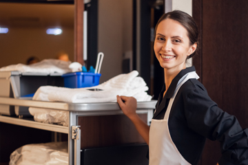 hotel maid standing by a trolley with towels and cleaning products, she is looking at the camera and smiling