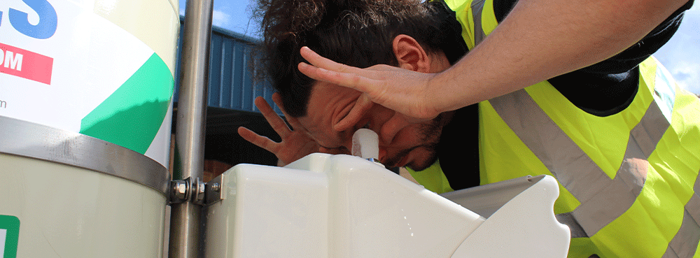A male worker wearing navy overalls holds his eyes open to rinse them with tepid water at a closed bowl eye wash station.