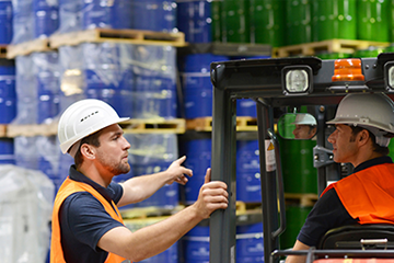 two workers in high vis, one in a forklift truck and the other directing in a chemical storage warehouse