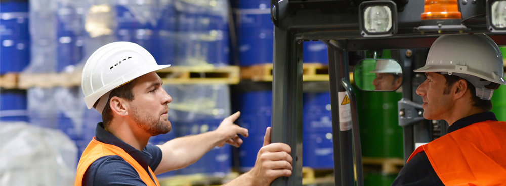 two workers in high vis, one in a forklift truck and the other directing in a chemical storage warehouse