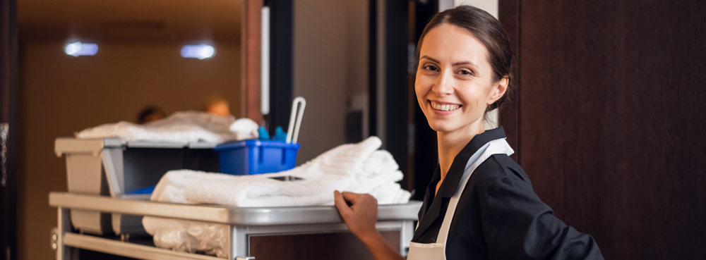 hotel maid standing by a trolley with towels and cleaning products. She is looking at the camera and smiling.